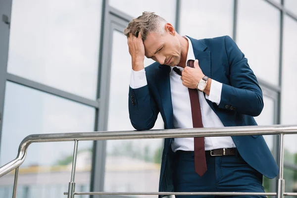 Upset businessman in suit standing outside and touching tie near building — Stock Photo