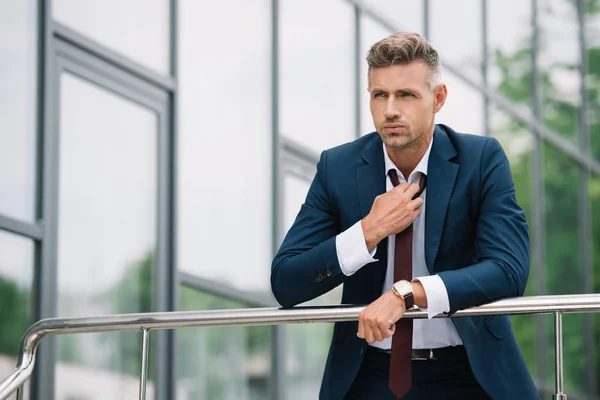 Handsome businessman in suit standing outside and touching tie near building — Stock Photo