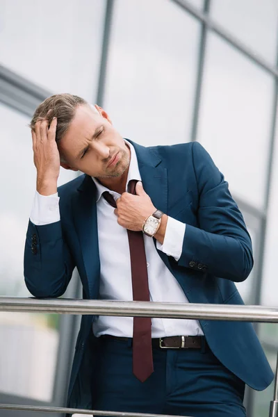 Molesto hombre de negocios en traje de pie fuera y tocando corbata cerca del edificio - foto de stock