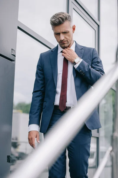 Selective focus of frustrated businessman in formal wear touching tie outside near building — Stock Photo