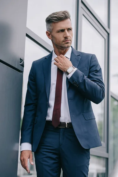 Serious businessman in formal wear touching tie outside near building — Stock Photo