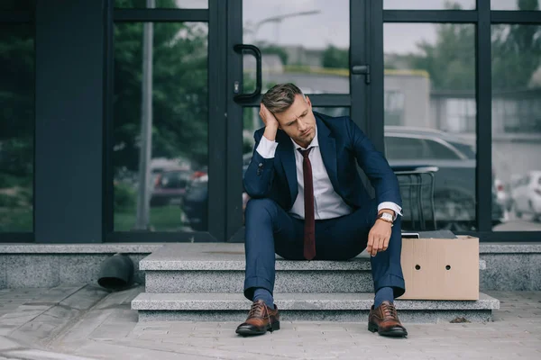Уволенный бизнесмен в костюме сидит на лестнице рядом с коробкой и трогает волосы — стоковое фото