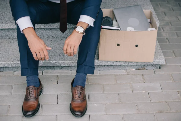 Vista ritagliata di licenziato uomo d'affari in giacca e cravatta seduto sulle scale vicino scatola di cartone — Foto stock