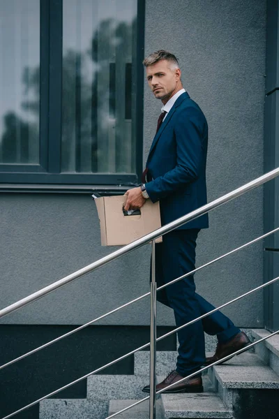 Уволенный бизнесмен, идущий по лестнице рядом со зданием и держащий коробку — стоковое фото