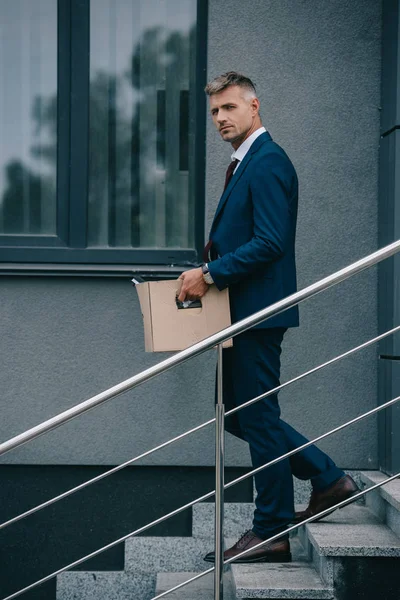 Hombre de negocios despedido caminando por las escaleras cerca de la construcción y la celebración de caja de cartón - foto de stock