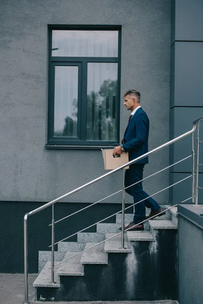 Vista laterale dell'uomo d'affari licenziato che cammina sulle scale vicino alla costruzione e tiene la scatola di cartone — Foto stock