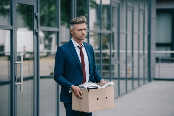 Sad and fired businessman standing near building and holding carton box — Stock Photo