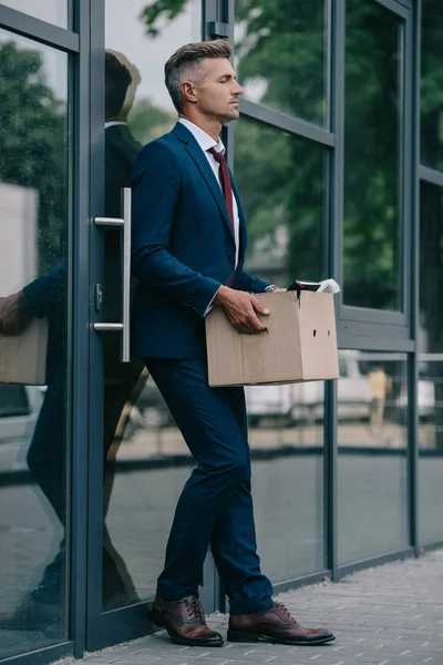 Upset and fired businessman standing near building and holding carton box — Stock Photo