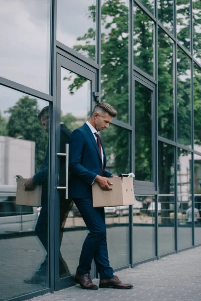 Upset and fired businessman standing near building and looking at carton box — Stock Photo