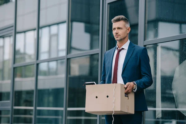 Dismissed businessman in suit standing near building with carton box — Stock Photo