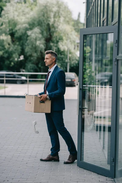 Fired man in suit walking near building with retro phone in carton box — Stock Photo