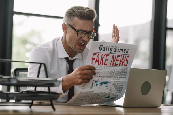 Selective focus of emotional businessman in glasses gesturing while reading newspaper with fake news — Stock Photo