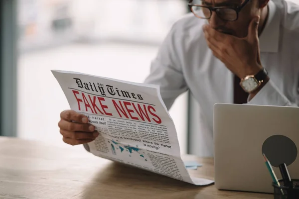 Choqué homme d'affaires dans des lunettes couvrant la bouche tout en lisant le journal avec de fausses nouvelles — Photo de stock