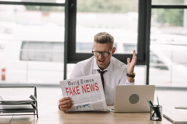 Расстроен бизнесмен в очках жестикулируя во время чтения газеты с фейковыми новостями — стоковое фото