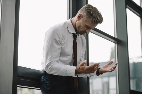 Molesto hombre de negocios en traje mirando las manos cerca de las ventanas — Stock Photo