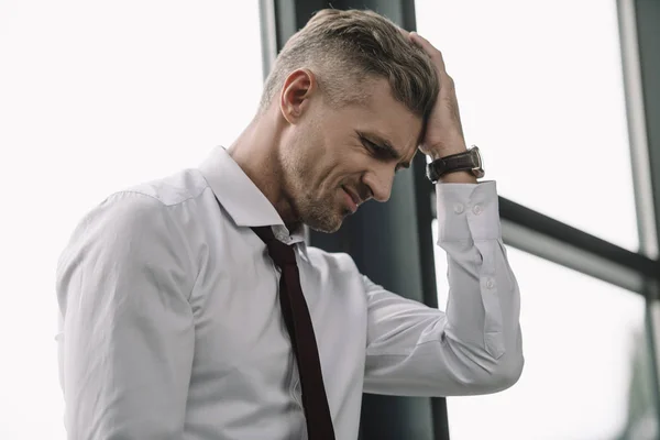 Расстроенный бизнесмен трогает волосы, стоя возле окон — стоковое фото