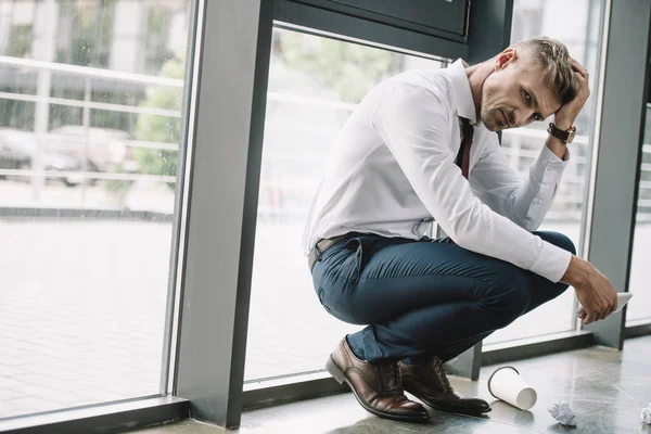 Frustrated businessman sitting near windows and holding smartphone — Stock Photo