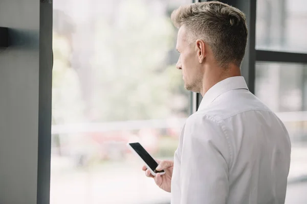 Guapo hombre de negocios sosteniendo teléfono inteligente con pantalla en blanco cerca de la ventana - foto de stock