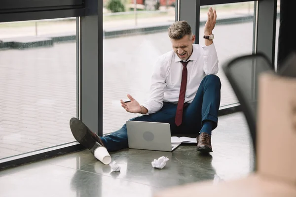 Вибірковий фокус незадоволеного чоловіка жестикулює, сидячи на підлозі і дивлячись на ноутбук — стокове фото