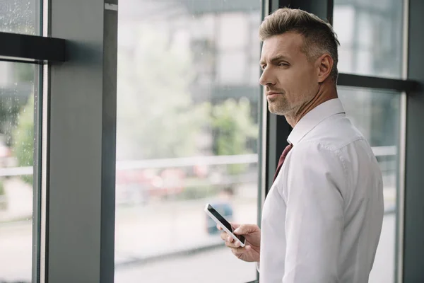 Homem bonito segurando smartphone com tela em branco perto da janela — Fotografia de Stock