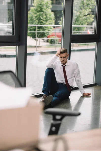 Избирательный фокус расстроенного бизнесмена, сидящего на полу рядом с окнами в офисе — стоковое фото