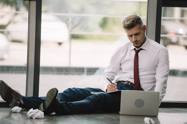 Selective focus of man writing in notebook near laptop while sitting on floor — Stock Photo