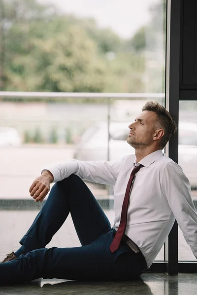 Hombre de negocios guapo sentado en el piso cerca de ventanas en la oficina - foto de stock