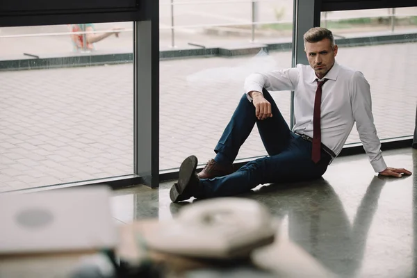 Selektiver Fokus eines verärgerten Geschäftsmannes, der auf dem Fußboden in der Nähe von Fenstern und Arbeitsplatz sitzt — Stockfoto