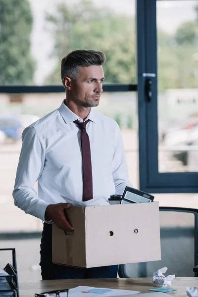 Красивый бизнесмен в костюме держит коробку рядом с рабочим местом — стоковое фото