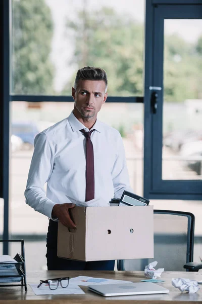 Serious businessman holding box while standing near workplace — Stock Photo