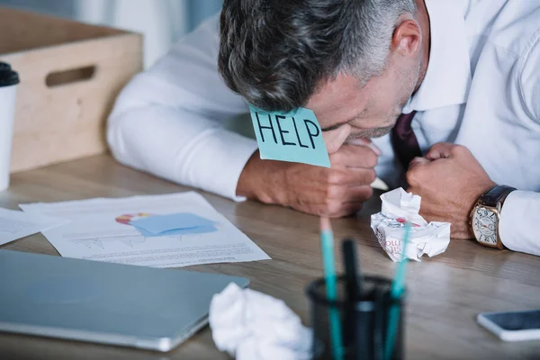 Homem de negócios com nota pegajosa na testa perto de mesa de bolas de papel — Fotografia de Stock