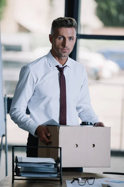 Entlassener Geschäftsmann hält Schachtel in der Nähe seines Arbeitsplatzes und schaut in die Kamera — Stockfoto