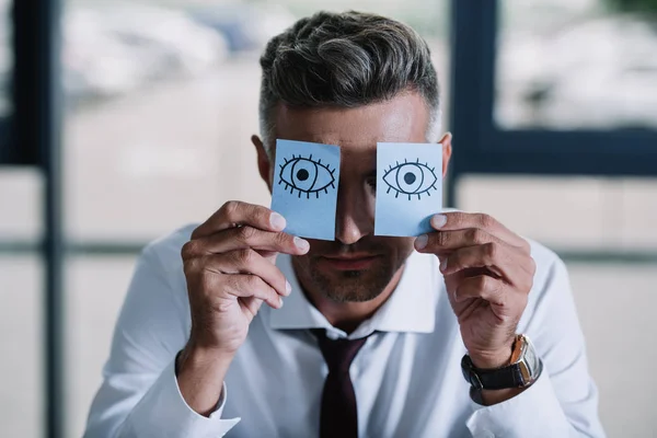 Hombre de negocios en traje sosteniendo notas adhesivas con los ojos dibujados cerca de la cara - foto de stock