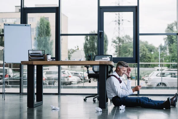 Hombre de negocios estresado sentado en el piso cerca de la mesa en la oficina moderna - foto de stock