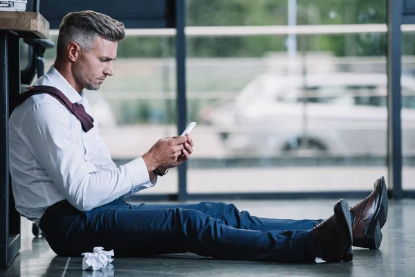 Seitenansicht eines Geschäftsmannes, der auf dem Boden sitzt und sein Smartphone benutzt — Stockfoto