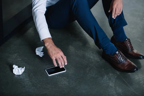 Vista recortada del hombre de negocios sentado en el piso cerca de teléfono inteligente con pantalla en blanco - foto de stock