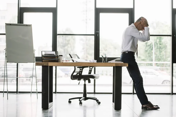 Guapo hombre de negocios tocando la cabeza mientras está de pie cerca de la mesa en la oficina moderna - foto de stock