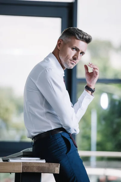 Handsome businessman standing near wooden table and looking at camera — Stock Photo