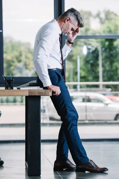 Bouleversé homme debout près de la table en bois et toucher la tête dans le bureau — Photo de stock