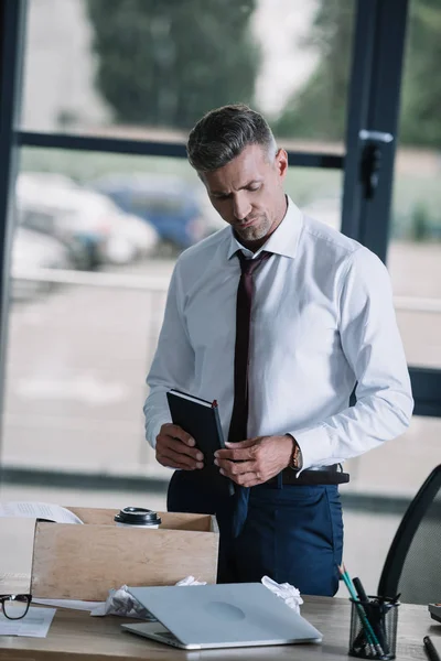 Unzufriedener Geschäftsmann hält Notizbuch neben Schachtel auf Tisch — Stockfoto