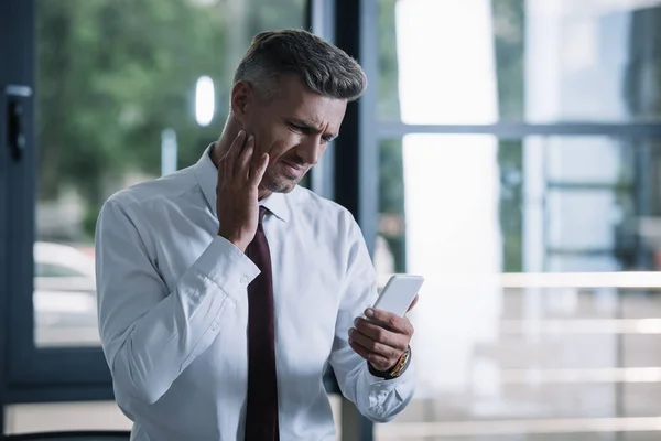 Molesto hombre de negocios en traje mirando el teléfono inteligente en la oficina - foto de stock