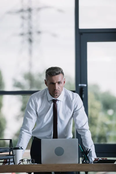 Серьезный бизнесмен в формальной одежде стоит у стола и смотрит в камеру — стоковое фото
