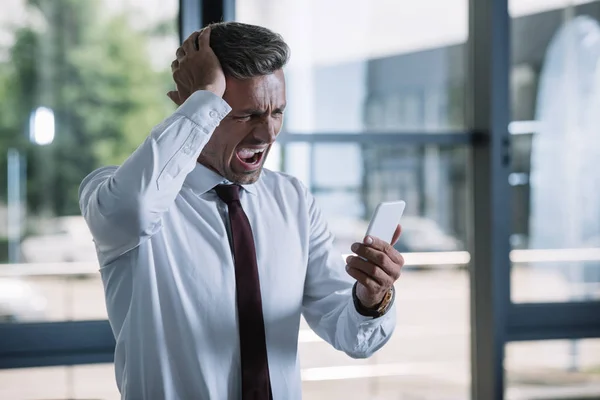 Molesto hombre de negocios mirando teléfono inteligente y gritando en la oficina - foto de stock