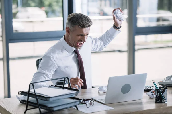 Selective focus of angry man holding crumpled paper ball and looking at laptop — Stock Photo