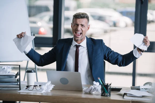 Hombre enojado sosteniendo papeles arrugados cerca de la computadora portátil en la mesa - foto de stock