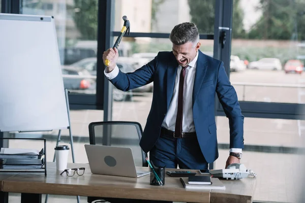 Сердитый бизнесмен держит молоток возле ноутбука в офисе — стоковое фото