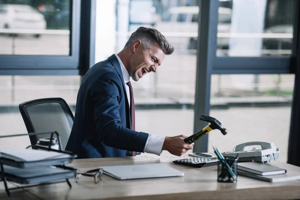 Enfoque selectivo de hombre de negocios enojado sosteniendo martillo cerca del teléfono retro en la oficina - foto de stock