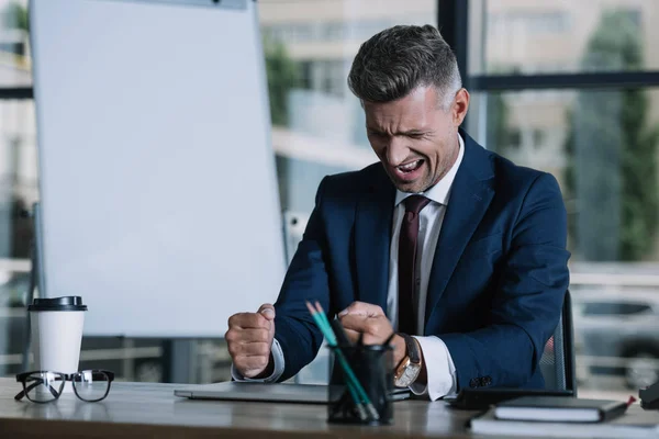 Enfoque selectivo del hombre enojado sentado cerca de la computadora portátil con los puños cerrados en la oficina - foto de stock