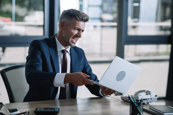 Bouleversé homme tenant ordinateur portable près du smartphone dans le bureau — Photo de stock