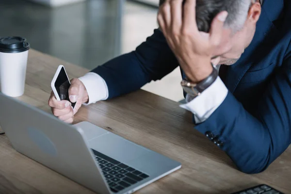 Вибірковий фокус засмученого чоловіка в костюмі, що тримає смартфон біля ноутбука — стокове фото